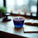 נר סויה ריחני בכוס כחולה, מואר ליד חלון עם טיפות מים, ספר פתוח לידו.